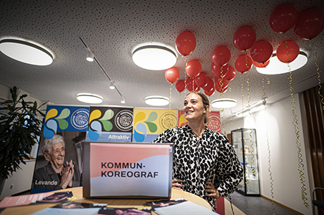 Malin Haglund står framför publik. Framför henne står en dator med texten Kommunkoreograf, i bakgrunden syns flera röda ballonger