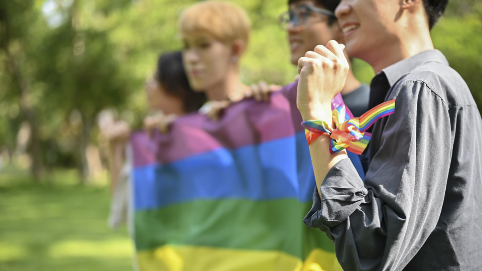 Fyra unga personer håller i en regnbågsflagga. Personen närmast kameran har ett regnbågsband runt handleden.