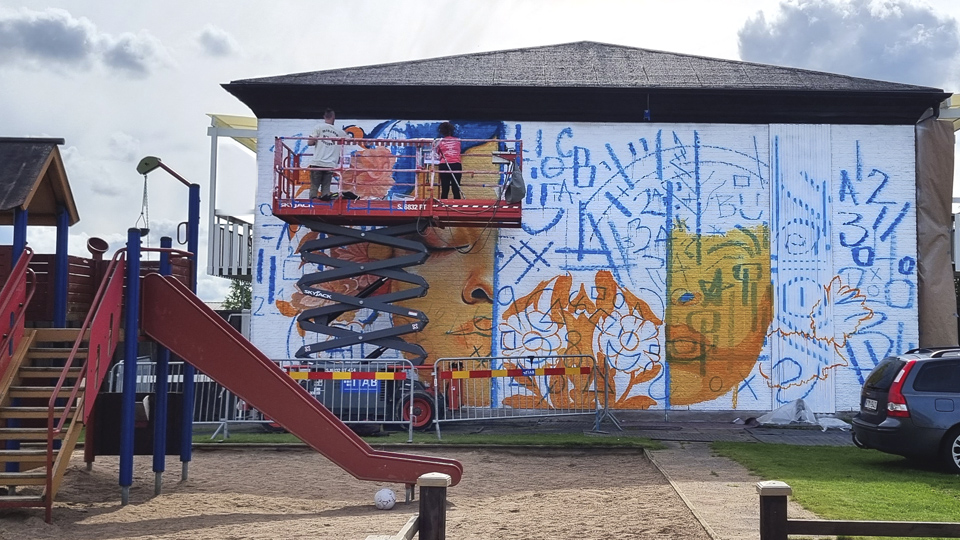 Två konstnärer på en skylift målar en gavelvägg på ett flerfamiljshus i Vargön.