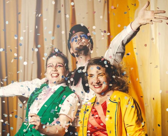 Tre cirkusartister i glada färger ståendes i ett konfettiregn