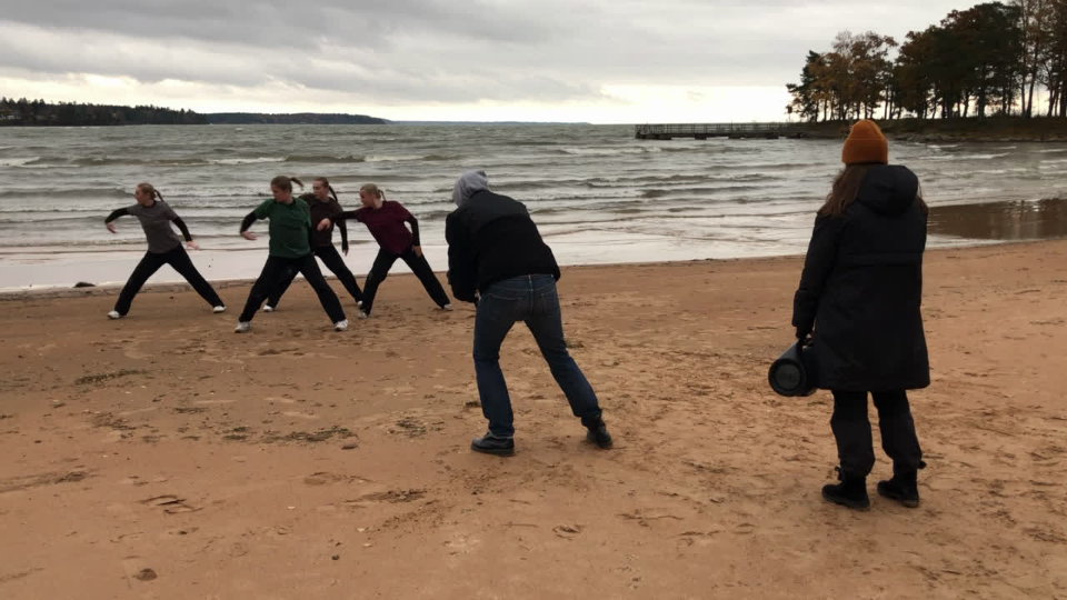 En bild på en grupp människor på en strand med Vänern i bakgrunden. Närmast vattnet dansar fyra ungdomar. Framför dem står en person som filmar dansen och bakom filmaren står en person med en högtalare. 