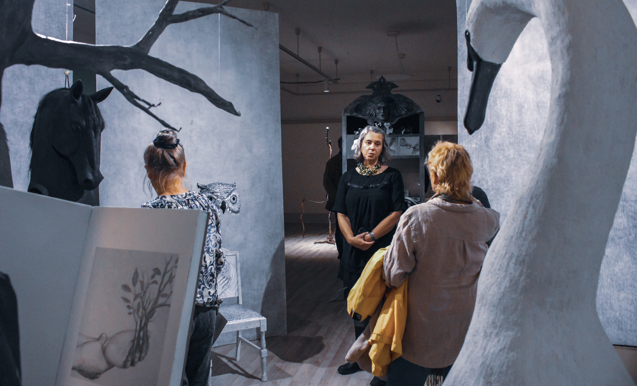 Visning i konsthallen. Besökare får berättat för sig om Vanna Bowles installation. Fåglar och hästhuvuden i gråa toner syns i bild.