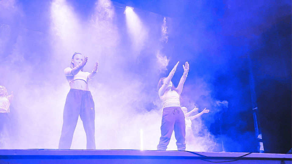 Fyra dansare som står på en scen. Scenbelysningen är grön och det är rök i bakgrunden. Dansarna har på sig svarta byxor och ljusblåa tröjor. De har händerna utsträcka rakt fram.