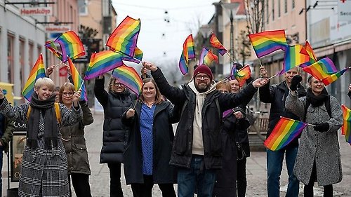 Flera personer står på gågatan i Vänersborg och viftar med regnbågsflaggor.