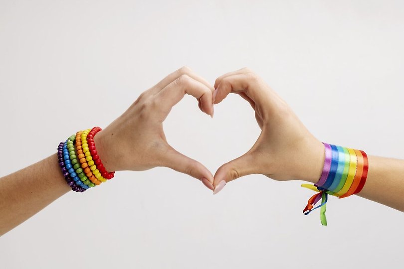 Två händer med regnbågsfärgade band runt handlederna bildar ett gemensamt hjärta
