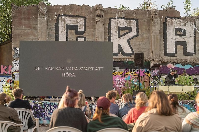 Miljöbild över arrangemanget: bioskärm, DJ, graffitiklädda betongväggar och sittande publik.